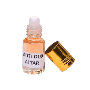 May include: A small, clear glass bottle with a roll-on applicator, containing a light orange liquid. The label reads "MITTI OUD ATTAR." A gold-coloured cap is next to the bottle. The product is likely a fragrance or essential oil.