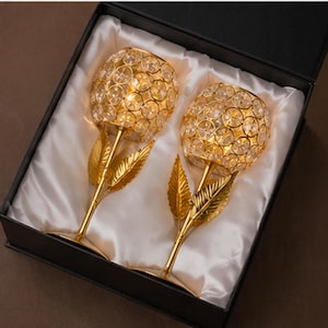 Peut inclure: Deux bougeoirs dorés en forme de rose avec des ornements en forme de cristal, présentés dans un coffret noir avec une doublure en satin blanc. Les bougeoirs ont une tige et un motif de feuilles.