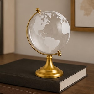 Op de afbeelding: Een globe van helder glas met matwitte continenten en dunne, gegraveerde lengte- en breedtegraden. De globe wordt ondersteund door een goudkleurige metalen standaard met een ronde voet en een gebogen arm die de bol op zijn plaats houdt.