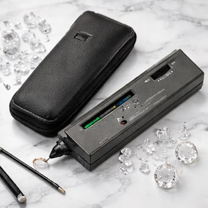May include: A black diamond tester with a leather case and a collection of clear, faceted gemstones. The tester is rectangular with a digital display and a probe. The gemstones are scattered around the tester.