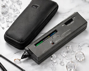 May include: A black diamond tester with a leather case and a collection of clear, faceted gemstones. The tester is rectangular with a digital display and a probe. The gemstones are scattered around the tester.