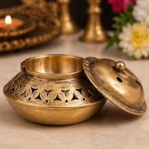 Può includere: Un bruciatore di incenso decorativo in ottone con coperchio. Il bruciatore ha una forma a ciotola rotonda e aperta con intricati motivi ritagliati sui lati. Il coperchio è leggermente socchiuso, rivelando l'interno. Il colore generale è un caldo oro antico.