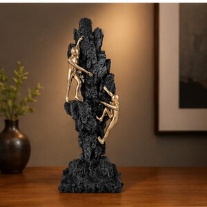 May include: A black sculpture of a rock formation with three gold-coloured figures climbing. The figures are in various climbing poses, and the rock has a textured surface. The sculpture is on a wooden surface.