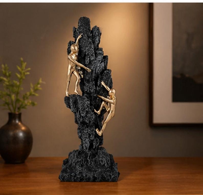 May include: A black sculpture of a rock formation with three gold-coloured figures climbing. The figures are in various climbing poses, and the rock has a textured surface. The sculpture is on a wooden surface.