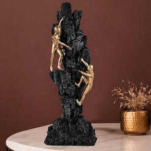 May include: A black and gold sculpture of two figures climbing a textured rock formation. The figures are gold, and the rock is black. The sculpture is on a white marble table.