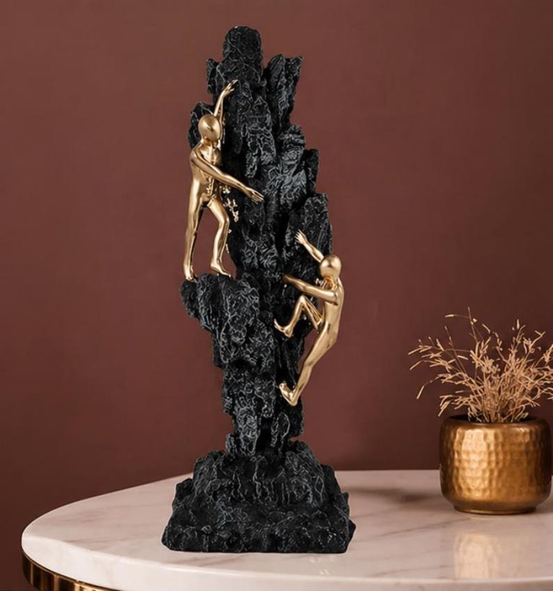 May include: A black and gold sculpture of two figures climbing a textured rock formation. The figures are gold, and the rock is black. The sculpture is on a white marble table.