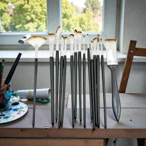 May include: A collection of artist paint brushes with black handles and white bristles, arranged on a wooden table. The brushes vary in size and shape, including a fan brush. A palette and paint tubes are visible in the background.