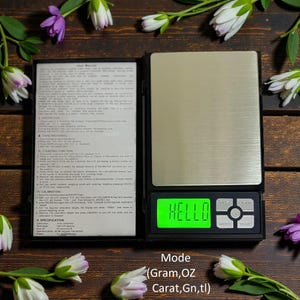 May include: A digital pocket scale with a rectangular stainless steel weighing surface. The display shows "HELLO" in green. The scale has buttons for mode, tare, and clear. The text "Mode (Gram, OZ Carat, Gn, tl)" is also visible.