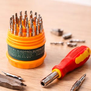 Puede incluir: Un juego de puntas de destornillador naranja y verde con un destornillador con mango rojo y amarillo. El juego incluye una variedad de puntas plateadas y está diseñado para trabajos eléctricos. Las herramientas están dispuestas sobre una superficie de madera.