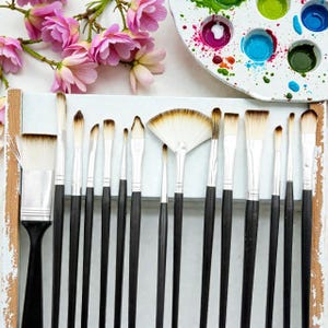 May include: A collection of artist paint brushes with black handles and white bristles, arranged in a white wooden holder. A fan brush is in the center. Pink flowers and a paint palette with colorful paint are in the background.