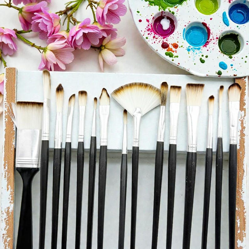May include: A collection of artist paint brushes with black handles and white bristles, arranged in a white wooden holder. A fan brush is in the center. Pink flowers and a paint palette with colorful paint are in the background.