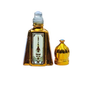 Puede incluir: Un frasco de perfume dorado con un diseño decorativo y una tapa separada. El frasco tiene forma rectangular con un panel vertical detallado. La tapa tiene forma de cúpula con una pequeña gema azul en la parte superior.