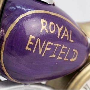 Könnte beinhalten: Nahaufnahme eines violetten Dekorationsobjekts mit den goldfarbenen Worten "ROYAL ENFIELD". Das Objekt hat eine abgerundete Form und scheint Teil eines größeren Dekorationsobjekts zu sein, möglicherweise einer Uhr oder Skulptur.
