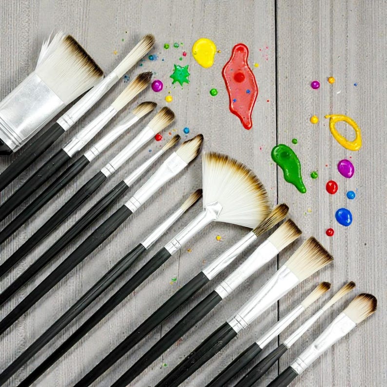 May include: An assortment of paint brushes with black handles and silver ferrules, arranged diagonally on a gray wooden surface. Splatters of yellow, red, green, purple, and blue paint are scattered around the brushes, suggesting artistic activity.