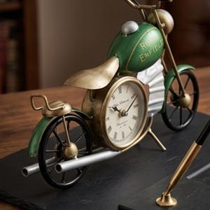 Könnte beinhalten: Eine dekorative Motorraduhr in Grün und Gold. Das Zifferblatt ist rund mit römischen Ziffern. Auf dem Tank steht "Royal Enfield". Das Motorrad hat schwarze Räder und einen silbernen Auspuff. Die Uhr steht auf einer schwarzen Oberfläche.