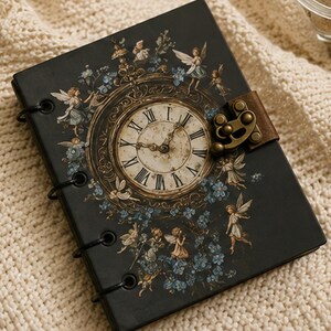 May include: A black rectangular journal featuring a central clock face with Roman numerals, surrounded by illustrations of small winged figures and blue flowers. The cover includes a brown leather strap with a metallic bronze-toned latch closure.
