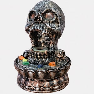 Puede incluir: Una fuente decorativa con forma de calavera con un acabado metálico texturizado. La calavera tiene detalles y un pequeño cuenco en la boca. La base está adornada con piedras de colores. Un artículo de decoración para el hogar de temática gótica.