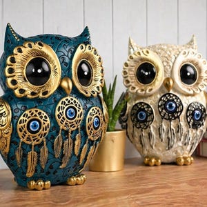 Op de afbeelding: Twee decoratieve uilbeeldjes. Eén is teal met gouden accenten, de andere is gebroken wit met gouden details. Beide hebben grote zwarte ogen en ingewikkelde dromenvanger-ontwerpen met blauwe oogsymbolen.