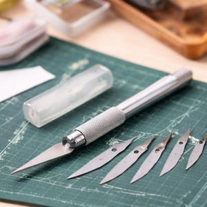 May include: A silver craft knife with a textured handle and a set of replacement blades. The blades are sharp and pointed, and the knife is on a green cutting mat. A clear plastic case is also present.