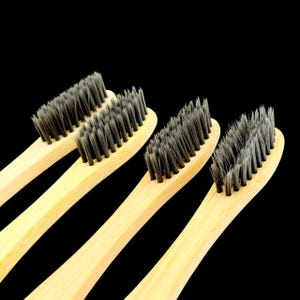 以下が含まれることがあります： 黒い背景に、灰色の毛が付いた4本の竹製歯ブラシが斜めに配置されています。ハンドルは明るい自然な木の色です。毛は密に詰まっており、柔らかく見えます。