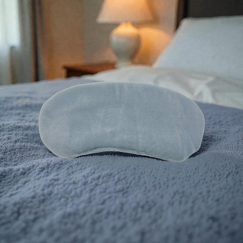 Pu&ograve; includere: Una mascherina per dormire azzurra realizzata in morbido materiale felpato. La mascherina, di forma ovale, &egrave; appoggiata su una coperta blu strutturata. Lo sfondo include un letto con lenzuola bianche e una lampada.