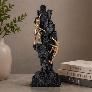 May include: A black resin sculpture depicting a mountain with two gold figures climbing. This decorative piece is likely for a home or office. The figures are in the process of climbing the mountain.