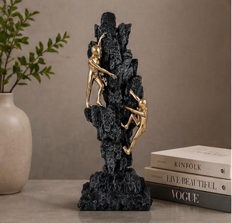 May include: A black resin sculpture depicting a mountain with two gold figures climbing. This decorative piece is likely for a home or office. The figures are in the process of climbing the mountain.