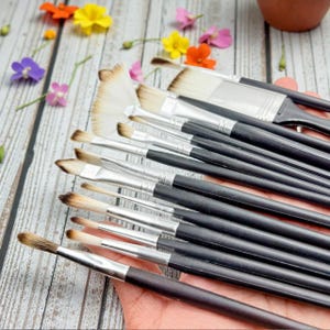 May include: A collection of paint brushes with black handles and silver ferrules. The bristles vary in shape and size, including a fan brush. Colorful flowers are scattered around the brushes, suggesting an art or craft theme.