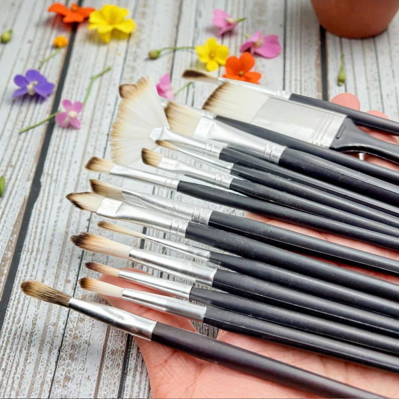 May include: A collection of paint brushes with black handles and silver ferrules. The bristles vary in shape and size, including a fan brush. Colorful flowers are scattered around the brushes, suggesting an art or craft theme.