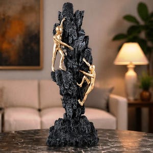 May include: A decorative sculpture featuring two gold figures climbing a textured black rock formation. The figures are in mid-climb, reaching upwards. A unique home decor item.