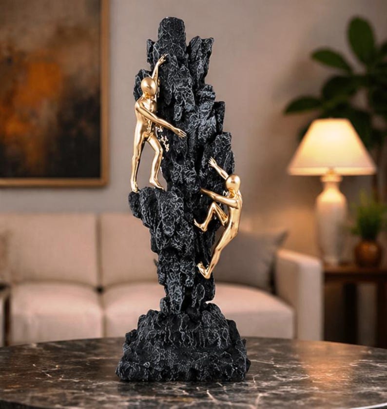 May include: A decorative sculpture featuring two gold figures climbing a textured black rock formation. The figures are in mid-climb, reaching upwards. A unique home decor item.