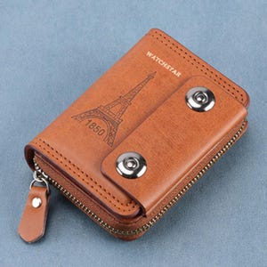 May include: A brown leather key wallet with a zipper closure and snap closure. The front features an embossed Eiffel Tower design with the text "1850" and "WATCHSTAR". The wallet is rectangular and has a textured surface.