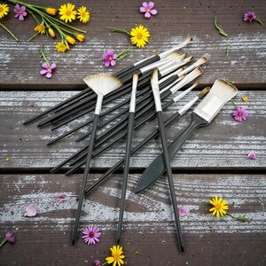 May include: A collection of paint brushes with black handles and silver ferrules, arranged on a weathered wooden surface. The brushes vary in size and shape, including a fan brush and a wide brush. Yellow and purple flowers are scattered around the brushes.