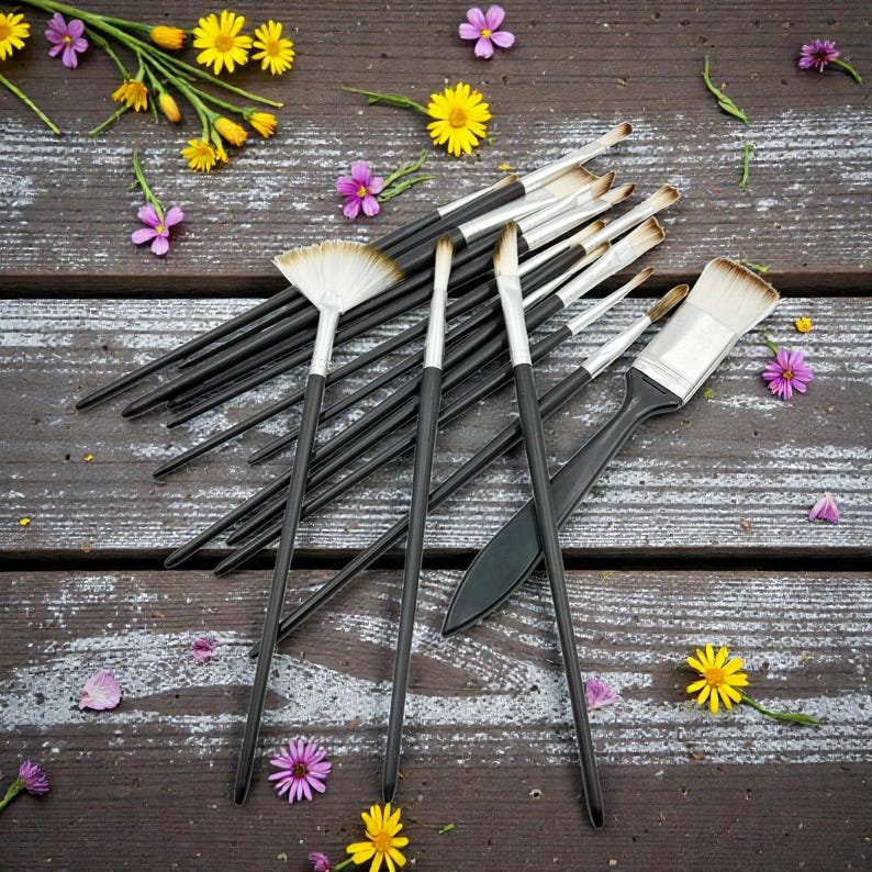 May include: A collection of paint brushes with black handles and silver ferrules, arranged on a weathered wooden surface. The brushes vary in size and shape, including a fan brush and a wide brush. Yellow and purple flowers are scattered around the brushes.