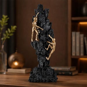 May include: A black sculpture depicting a rock formation with two gold figures climbing. The figures are ascending the textured, dark-coloured rock. The sculpture is a decorative item, suitable for display.