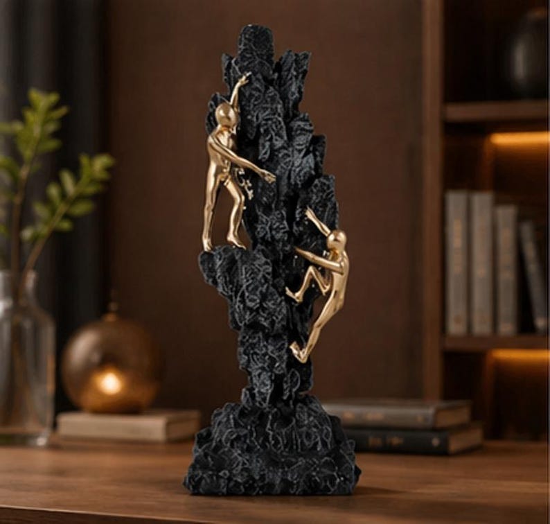 May include: A black sculpture depicting a rock formation with two gold figures climbing. The figures are ascending the textured, dark-coloured rock. The sculpture is a decorative item, suitable for display.