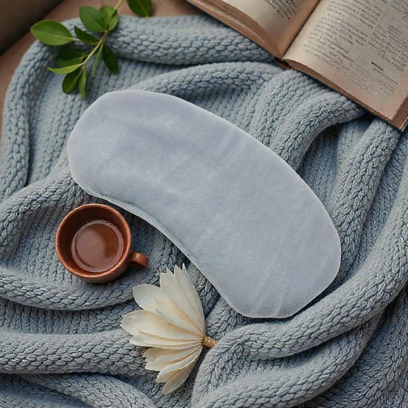 Pu&ograve; includere: Una mascherina per gli occhi azzurra &egrave; appoggiata su una coperta di lana grigia, accanto a una piccola tazza marrone e un fiore bianco. Un libro aperto &egrave; visibile sullo sfondo. La mascherina &egrave; realizzata in un materiale morbido e vellutato.