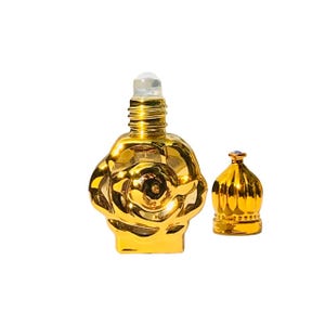 Puede incluir: Un frasco de perfume dorado con diseño de rosa, junto a un tapón a juego. El frasco tiene un aplicador de bola rodante y una superficie brillante y reflectante. El tapón es una pieza decorativa pequeña con una joya en la parte superior.