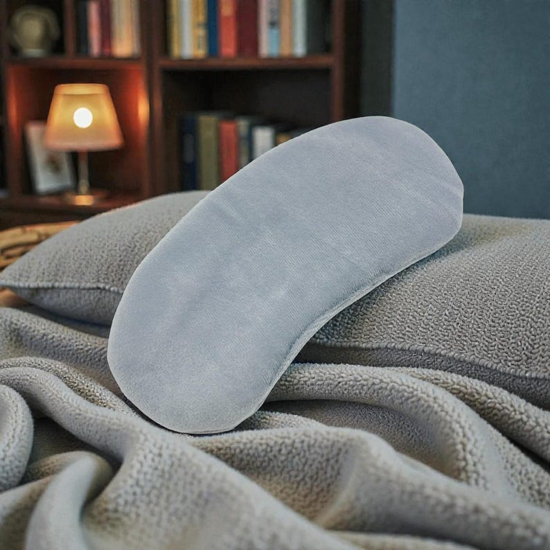 Pu&ograve; includere: Un cuscino curvo grigio chiaro poggia su una coperta e un cuscino grigi strutturati. Il cuscino ha un aspetto morbido e soffice. Lo sfondo include una libreria, una lampada e una parete blu scuro, creando un'atmosfera accogliente e rilassante.