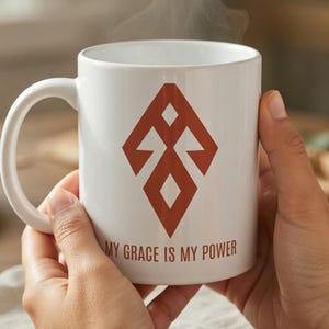 Puede incluir: Taza de cerámica blanca con un diseño geométrico marrón rojizo y el texto "MY GRACE IS MY POWER". Sale vapor de la taza, lo que sugiere una bebida caliente. La taza se sostiene con ambas manos.