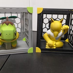 May include: Two toy figures, a green turtle-like creature and a yellow fox-like character, are displayed inside grey and black cages. The cages have yellow and green corner accents. The figures are on a black surface.