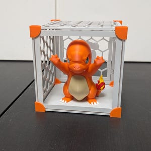 Peut inclure: Une figurine Charmander orange à l'intérieur d'une cage blanche et orange. La figurine a les bras levés et une flamme sur la queue. La cage est un cube avec des coins orange. Fond : mur blanc et surface noire.