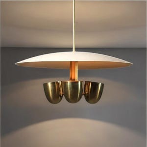 May include: A contemporary pendant light. It features a white, disc-shaped shade and three brass-coloured, cup-shaped lights. The fixture is suspended from a thin, gold-coloured rod.