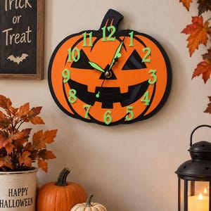 May include: A pumpkin-shaped wall clock featuring a bright orange face with a black jack-o'-lantern cutout design. The clock has raised, light green numbers from 1 to 12 and matching light green clock hands centered on the pumpkin's face.