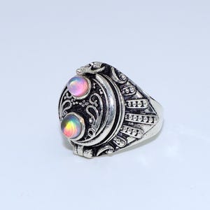 May include: A silver-toned ring with an oval, locket-style face. The face features two iridescent, round stones and ornate scrollwork. The ring has a detailed band with a textured design.