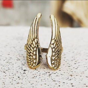 Puede incluir: Un anillo dorado con un diseño de dos alas abiertas. Las alas tienen texturas detalladas de plumas y están posicionadas para envolver un dedo. El anillo está sobre una superficie clara y moteada.