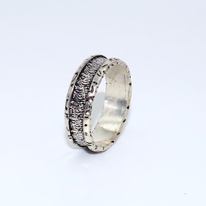 Puede incluir: Un anillo de plata con una banda texturizada. El anillo presenta un patrón detallado alrededor del centro, con un interior liso y pulido. El anillo está sobre un fondo blanco, resaltando su brillo metálico.