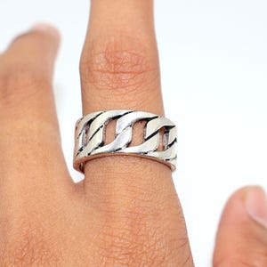 Puede incluir: Un anillo plateado con un diseño de eslabones de cadena. El anillo, que se lleva en un dedo, tiene una banda ancha con una serie de eslabones interconectados. El anillo está hecho de metal brillante y tiene un aspecto ligeramente desgastado.
