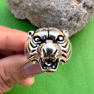Puede incluir: Un anillo plateado con forma de cabeza de tigre. El anillo presenta grabados detallados de la cara del tigre, incluyendo sus ojos, nariz, boca y rayas. El anillo se muestra sobre un fondo verde.