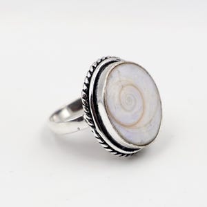 May include: A silver ring featuring a large, oval, white shell with a spiral pattern. The shell is set in a silver bezel with a rope-like border. The ring has a simple, polished band.
